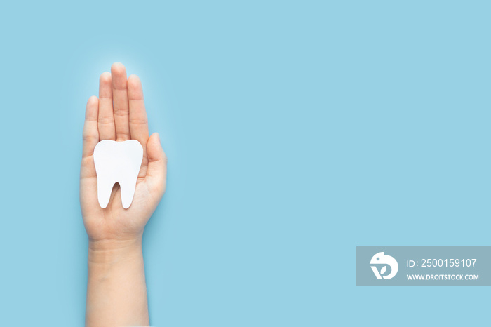 Woman hand holding healthy white teeth symbol on light blue background. Dental care, tooth protection and oral hygiene concept. Top view. Copy space.