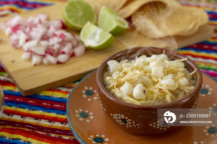 Pozole blanco con pollo, acompañado con rábanos, limones y tostadas en fondo de colores mexicanos