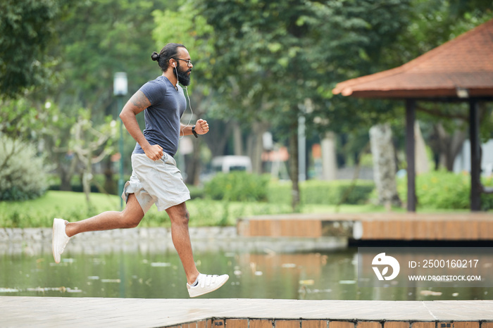 Side view of athletic bearded tattooed Indian man jogging in the morning outdoors and listening to music with earphones