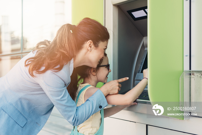 Young mother elegantly dressed with her daughter using ATM machine on city street.