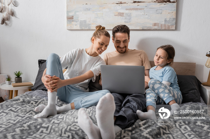 cheerful parents and happy kid watching movie on laptop in bedroom.