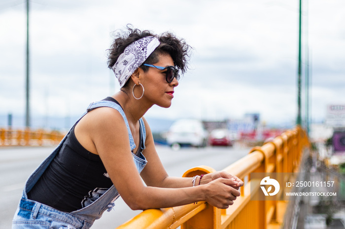 Mid adult afro mexican woman leaning on a bridge guard rail, looking at the distance, relaxing