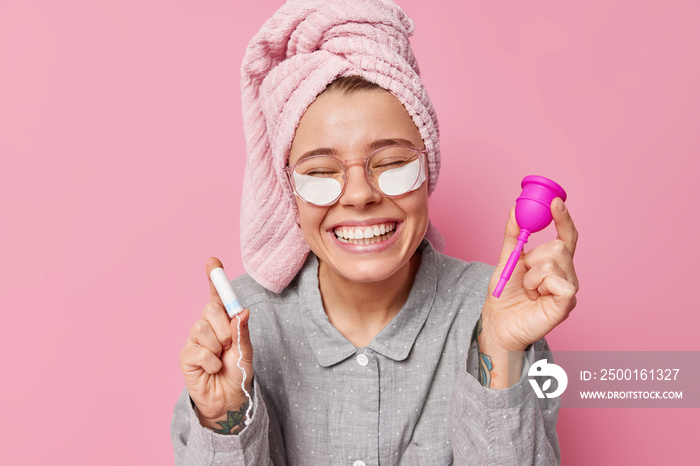 Positive young European woman wears comfortable pajama and wrapped towel on head holds tampon and menstruation cup during menses isolated over pink background applies beauty patches under eyes