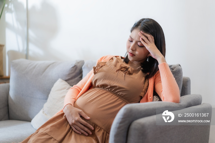 Young pregnant asian woman has suffering from headache sitting on sofa. Pregnancy symptoms