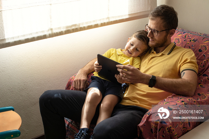 Papa con polo gialla gioca con il tablet insieme al figlio seduto nella poltrona di casa