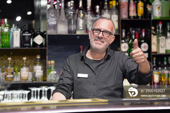 Professional elderly bartender in eyeglases show hand OK in workplace close-up. High level and quality service. The concept of service.