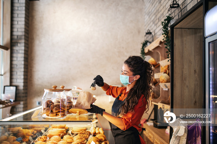 Beautiful young female worker with protective mask on face working in bakery.