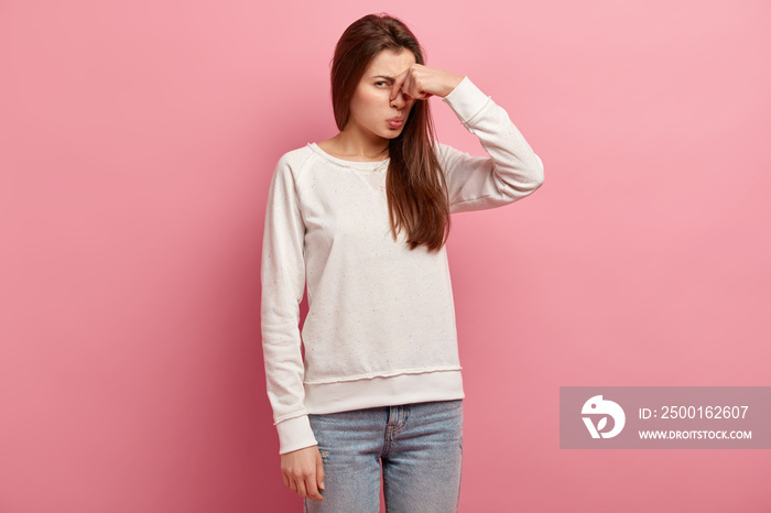 Displeased woman covers nose with hand, smells something awful, pinches nose, frowns in displeasure, sees pile of garbage, dressed in casual clothes, isolated over pink background. Bad odour.