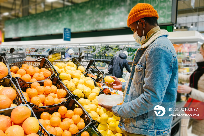 African-American man customer in warm jacket with disposable mask takes fresh lemon fruits from crate in supermarket side view