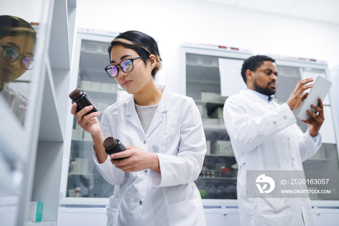 Serious concentrated young Asian female pharmacist with short hair standing at refrigerator with medications and finding necessary pills in medical storage room