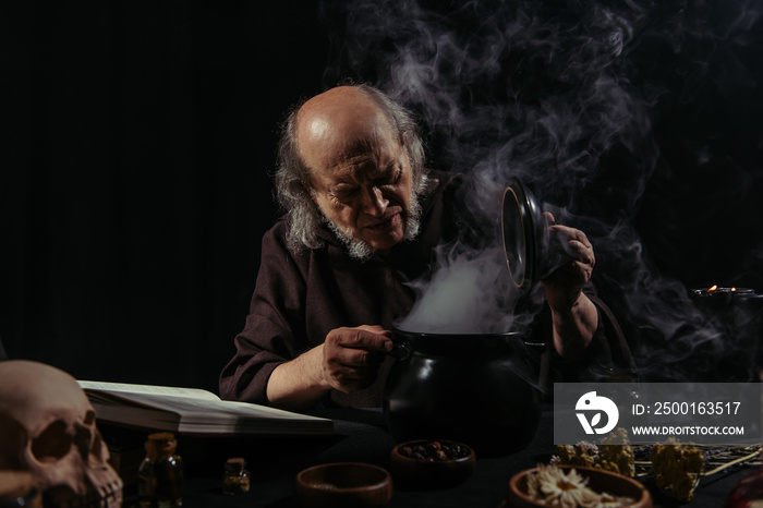 medieval alchemist near boiling pot and magic cookbook on black background.