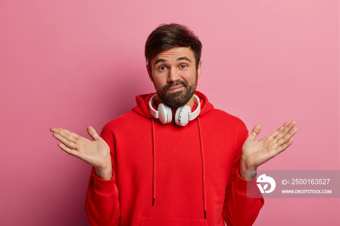 Indecisive millennial man with beard, shrugs shoulders, raises palms, dressed in red sweatshirt, uses stereo headphones, smirks problematic, cant understand anything, poses over rosy pastel wall