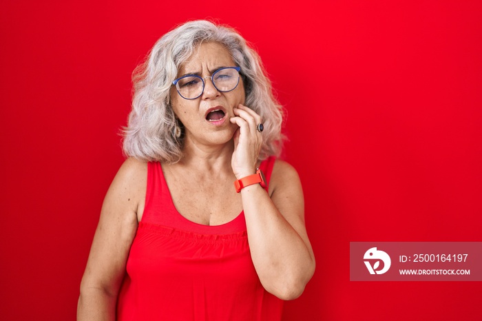 Middle age woman with grey hair standing over red background touching mouth with hand with painful expression because of toothache or dental illness on teeth. dentist