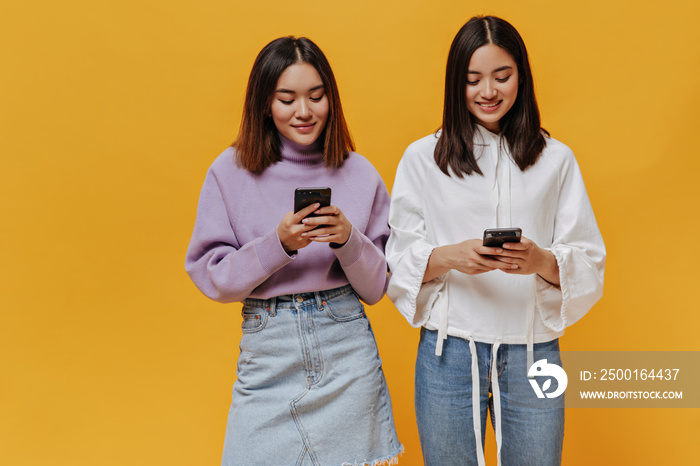 Joyful Asian girls hold phones on orange background. Brunette attractive women in sweatshirts and denim outfits messaging in cellphones on isolated.