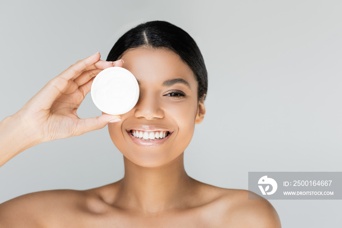 happy african american woman covering eye while holding container with face cream isolated on grey