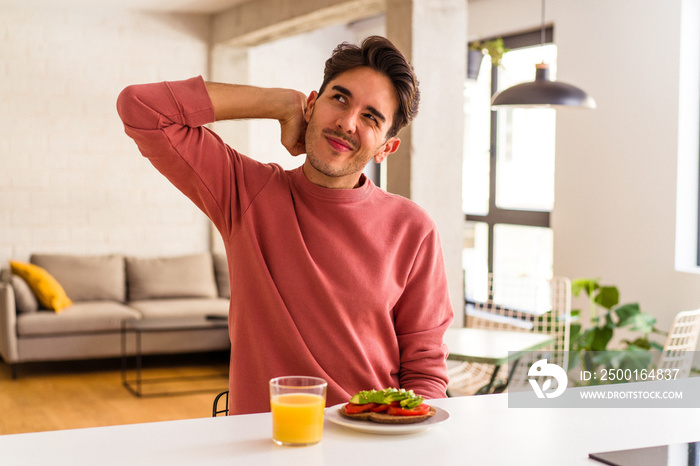 Young mixed race man having breakfast in his kitchen touching back of head, thinking and making a choice.