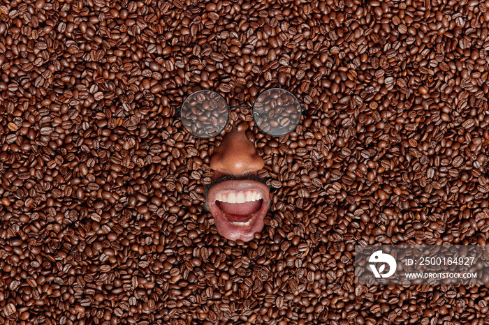 Happy dark skinned male model fully covered with brown coffee beans keeps mouth opened wears transparent glasses enjoys process of preparing favorite refreshing beverage. Top view. Creative shot