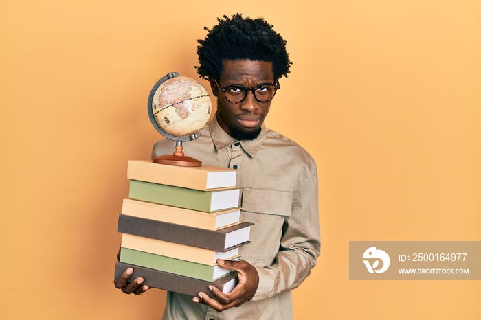 Young african american man holding a pile of books and vintage world ball skeptic and nervous, frowning upset because of problem. negative person.