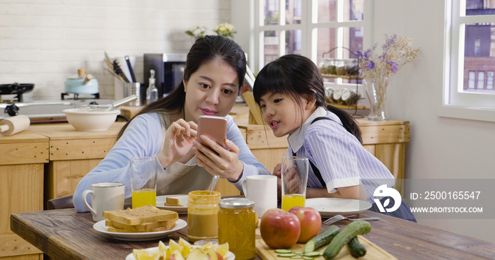 japanese cute little girl and beautiful young mom are sitting together at breakfast kitchen table using smart phone smiling. asian housewife with child talk chat online with dad work abroad cellphone