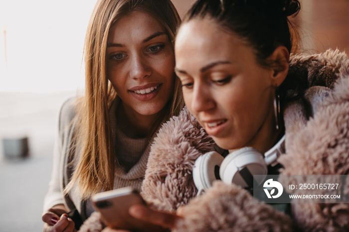 Latino woman with her friend is smiling happily using a smartphone and headphones in the city.