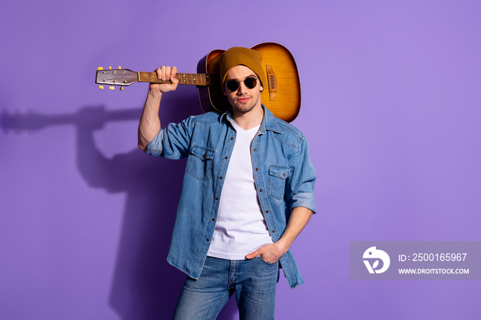 Photo of confident attractive handsome man wearing brown cap spectacles standing with guitar on his shoulder and hand in pocket isolated over purple vibrant color background