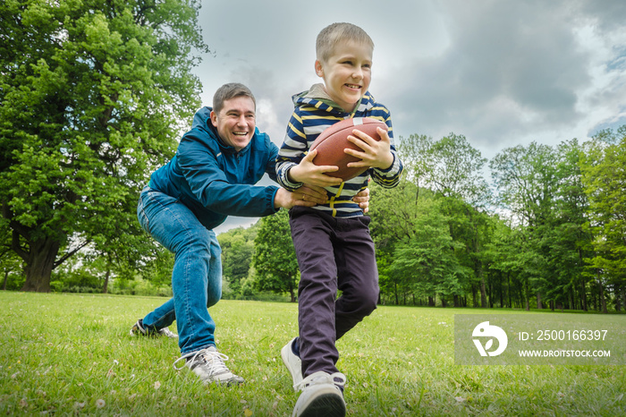 Father and son playing football, Father’s day, Playful Man teaching Boy rugby outdoors in sunny day at public park. Family sports weekend.