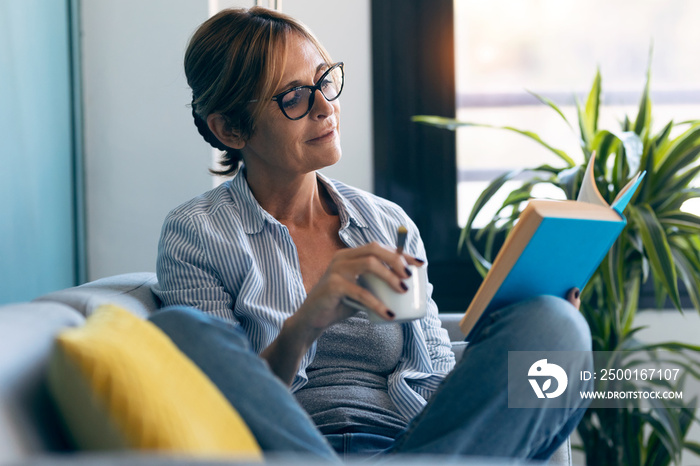 Mature beautiful woman reading a book while drinking a cup of coffee sitting on couch at home.