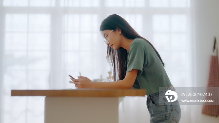 Happy young asian woman wearing glasses and relaxing at home. Asia female standing at counter kitchen and using mobile smartphone