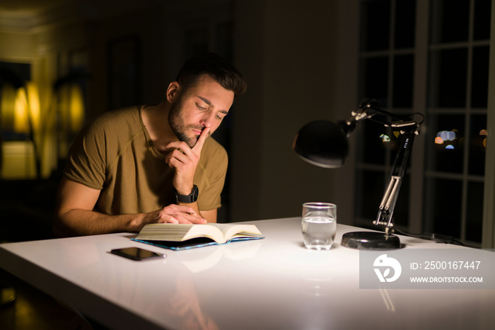 Young handsome man studying at home, reading a book at night