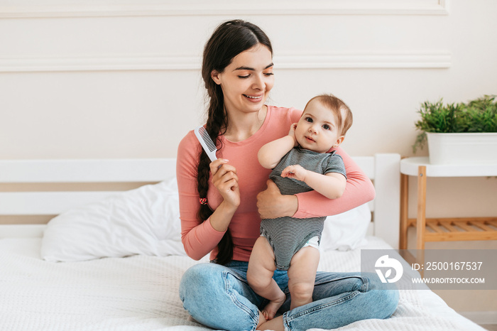 Happy mother combing hair of adorable baby girl, holding daughter on hands and smiling, little child looking at camera