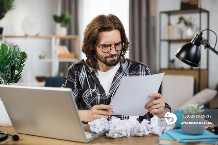 Angry caucasian guy sit at desk managing financial paperwork frustrated by bill amount. Unhappy young man feel distressed confused paying online, calculating expenses expenditures at home.