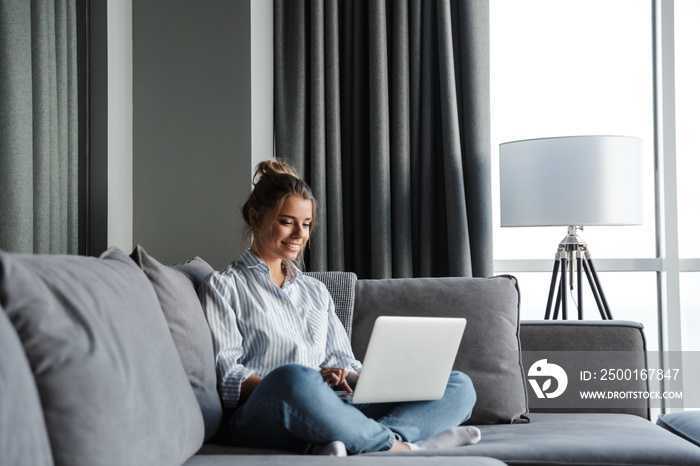 Image of joyful woman smiling and using laptop while sitting on couch