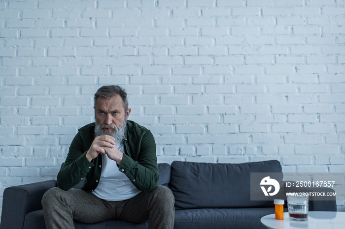 depressed senior man with grey hair sitting on couch near bottle with medication and glass of whiskey on coffee table.