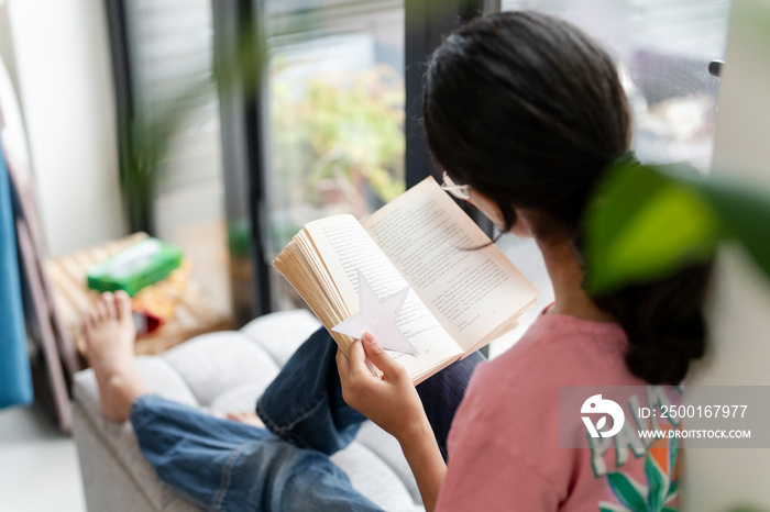 Girl sitting at hassock near window and reading book