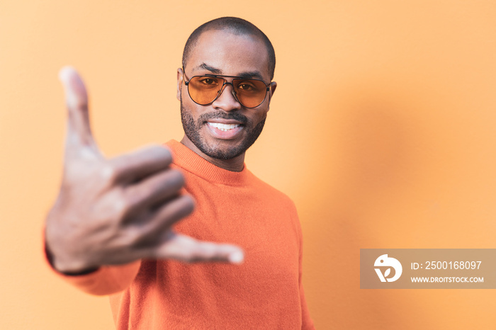 A dark-skinned adult man looks at the camera while making the Hawaiian shaka gesture of welcome. The man is wearing a shirt that is the same color as the background. Orange predominates.