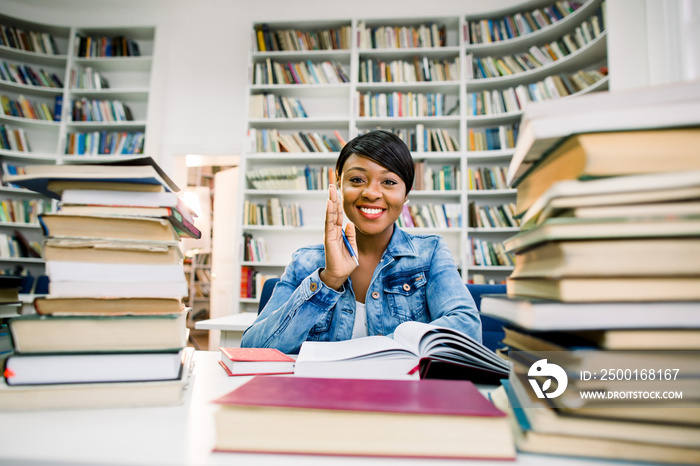 Portrait of young black african woman, reader, lady, literature lover, keeping arm on book sitting at a wooden table in the library touching cheek with hand looking to camera.