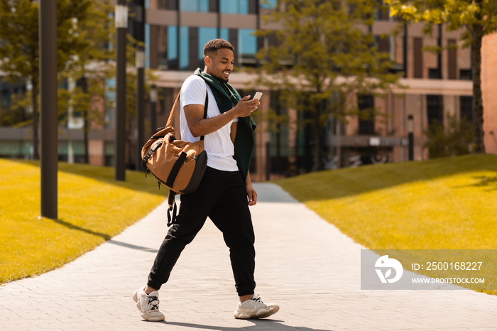 Young trendy black guy walking in the urban city area and texting on smartphone outdoors