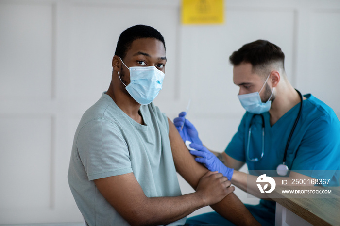 Health worker in surgical mask attending to black male patient, making coronavirus vaccine injection at clinic