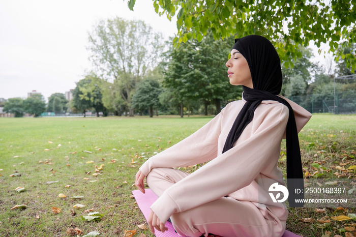 Woman in hijab and pink tracksuit meditating in park