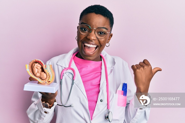 Young african american doctor woman holding anatomical model of female uterus with fetus pointing thumb up to the side smiling happy with open mouth