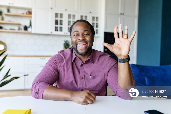 Glad to see you. African-American guy looks at the camera and waving hello, greeting online interloculor or participants of virtual meeting, involved in video call, virtual conference, webcam view