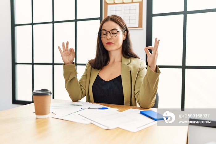 Young brunette woman wearing business style sitting on desk at office relax and smiling with eyes closed doing meditation gesture with fingers. yoga concept.