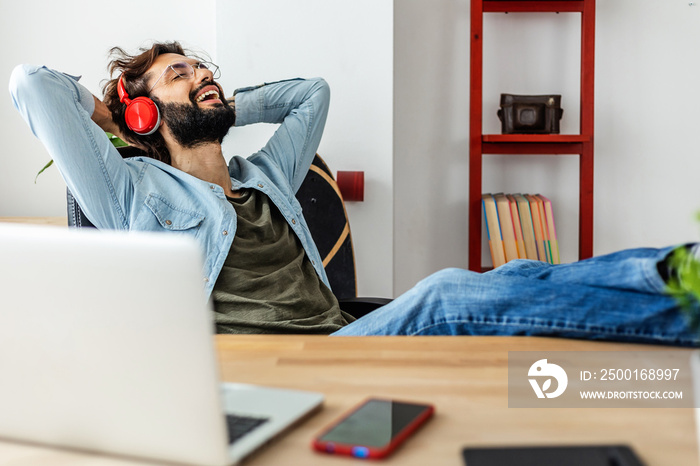 Smiling young adult hipster man relaxing at workplace. Happy freelancer male entrepreneur listening to music with headphones having a break from work.