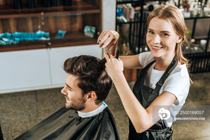 high angle view of hairstylist smiling at camera while cutting hair to handsome client in beauty salon