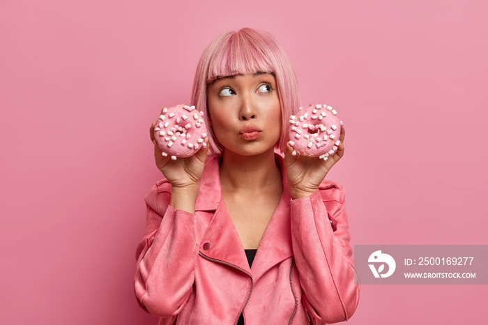 Photo of dreamy thoughtful Asian woman poses with two delicious donuts, has doubts to eat junk food or not, wears fashionable pink jacket, enjoys sweet dessert, stands indoor, rosy background