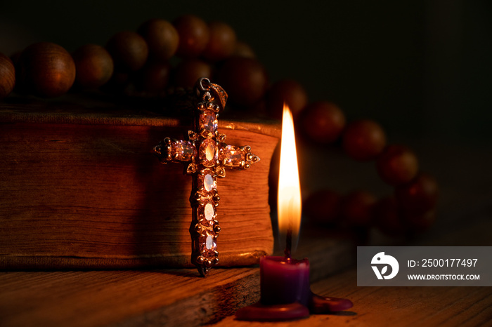 A bible cross and a wooden rosary lie on the table next to candles burning in the dark at home, prayer and faith, god