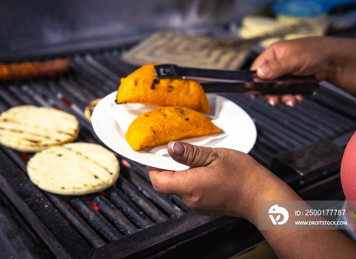 Hands serving two empanadas on a plate next to the grill with arepas and chorizo