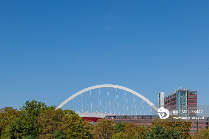 White arch steel beam structure and suspension cables of Lanxess Arena over treetop and Cologne cityscape in Germany. Arch superstructure beam with cable suspended support the roof and blue sky.