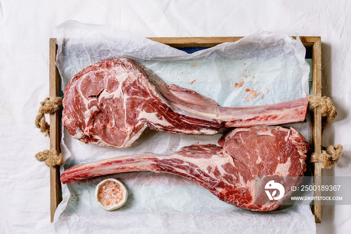 Raw uncooked black angus beef tomahawk steaks on bones served with pink salt on wooden tray with baking paper over white cloth as background. Top view, space.