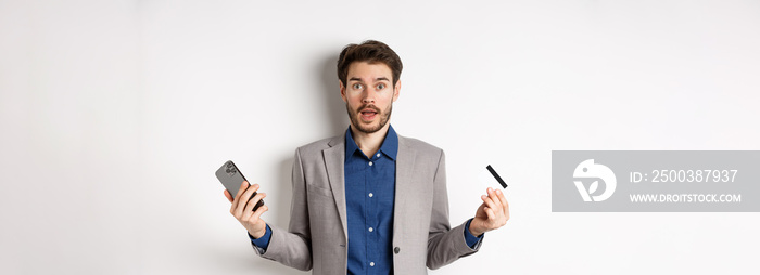 Online shopping. Confused man in suit spread hands sideways, holding plastic credit card with mobile